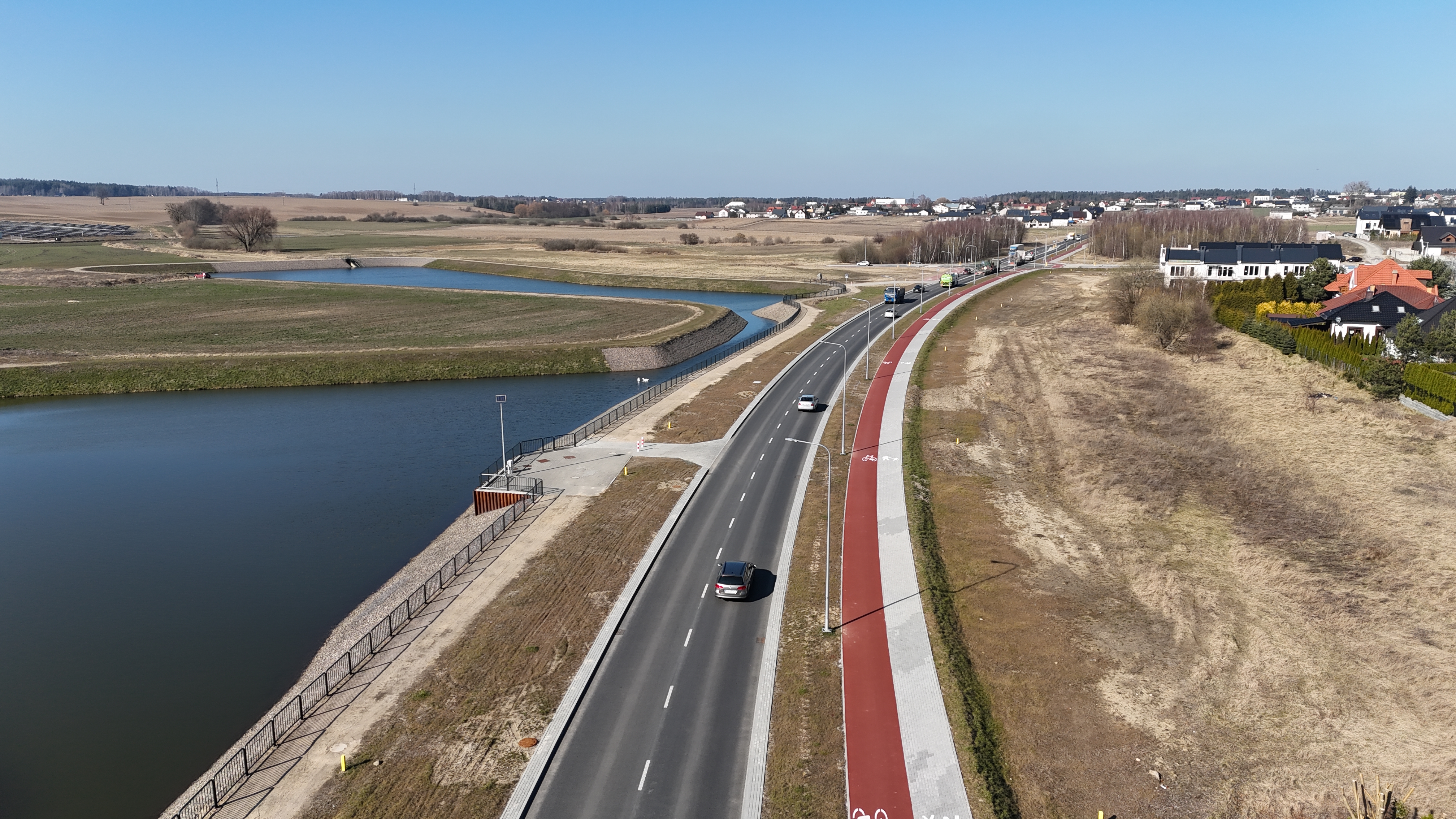 Widok na zachodnie obejście Chojnic w ciągu drogi wojewódzkiej nr 212 pod ruchem. W centrum kadru droga z pojazdami. Po prawej chodnik i ścieżka rowerowa oraz słupy oświetlenia ulicznego. W oddali widać zabudowania miasta, po lewej zbiorniki wodne (niezwiązane z inwestycją).