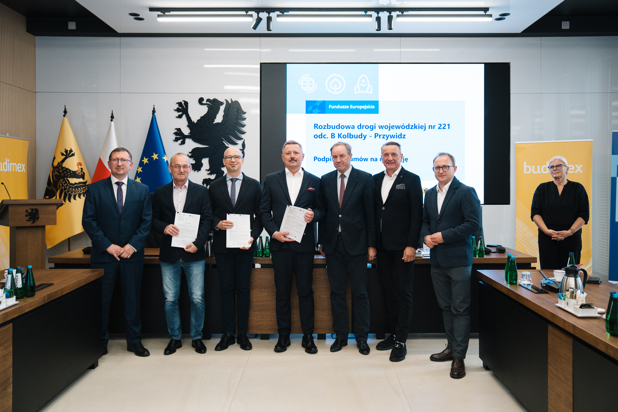 Grupa dziewięciu osób stoi razem, uśmiechając się do kamery, w sali konferencyjnej podczas oficjalnego podpisania umów. Trzech mężczyzn w środku trzyma w dłoniach dokumenty. W tle widać trzy flagi: województwa pomorskiego, Polski i Unii Europejskiej, a także ekran z napisem „Rozbudowa drogi wojewódzkiej nr 221 odc. B Kolbudy – Przywidz. Podpisanie umów na realizację”
